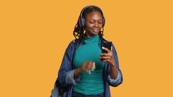 African american teenager talks with friends during conference using phone and headphones, studio background. Young BIPOC girl chatting with mates during internet call on cellphone, camera A video