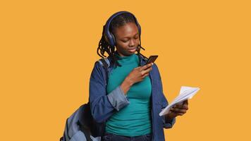 afrikansk amerikan kvinna tar bild av skola läxa till skicka till kollega, lyssnande musik, studio bakgrund. studerande fotografering papper med information behövs för examen, kamera en video