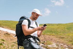 un barbado viajero con un mochila usos teléfono en el parte superior de un montaña. un turista con un mochila soportes en contra el antecedentes de un montaña foto