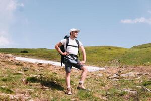 un barbado viajero con un mochila en el parte superior de un montaña. un turista con un mochila soportes en contra el antecedentes de un montaña foto