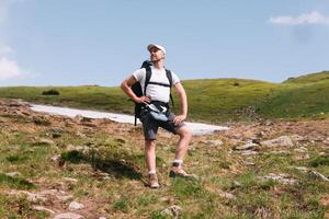 un barbado viajero con un mochila en el parte superior de un montaña. un turista con un mochila soportes en contra el antecedentes de un montaña foto