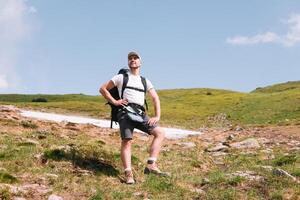 un barbado viajero con un mochila en el parte superior de un montaña. un turista con un mochila soportes en contra el antecedentes de un montaña foto