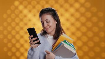 sonriente mujer leyendo mensajes en teléfono inteligente mientras participación apilar de libros, aislado terminado estudio antecedentes. alegre persona con pila de académico libros de texto en brazos mensajes de texto amigos en teléfono, cámara si video