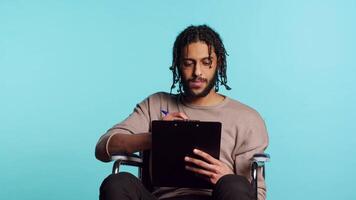 middle eastern man in wheelchair suffering from paraplegia holding clipboard in hand, using pen to write on paper. Person with disability completing forms, isolated over studio background, camera A video