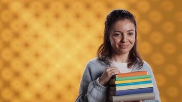 Portrait of smiling woman pointing towards empty copy spaces while holding pile of books. Radiant person with stack of novels in arms promoting reading, showing messages, studio background, camera A video