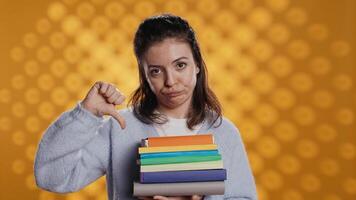 Portrait of woman with pouting expression holding pile of books, showing disproval of reading hobby. Sulky lady with stack of novels doing thumbs down hand gesturing, studio background, camera B video