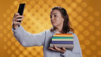 sorridente mulher segurando pilha do livros, desfrutando lendo passatempo, levando selfies com celular. alegre senhora com pilha do romances dentro braços fazendo As fotos com Móvel telefone, estúdio fundo, Câmera b video