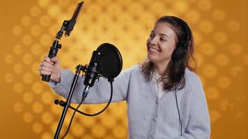 Joyous influencer filming herself reviewing book with selfie stick, isolated over studio background. Woman doing vlog with self portrait camera, talking with audience about novel, camera B video