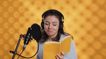 Happy narrator wearing headphones reading aloud from book into mic against yellow background. Joyous lady recording audiobook, creating engaging media content for listeners, camera A video