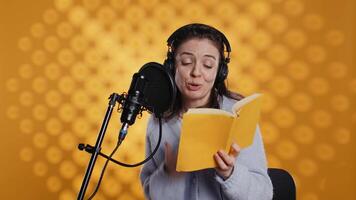 Happy narrator wearing headphones reading aloud from book into mic against yellow background. Joyous lady recording audiobook, creating engaging media content for listeners, camera B video