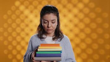 Portrait of smiling woman holding stack of books, doing salutation hand gesture, feeling optimistic. Jolly person with pile of novels raising arm to greet someone, studio background, camera B video