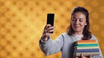 Reading enthusiast woman filming social media s with smartphone holding stack of books, studio background. Cheerful nerd with pile of textbooks in arms recording with selfie camera, camera A video