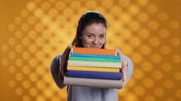 Portrait of friendly woman offering stack of textbooks useful for school exam, isolated over studio background. Merry person giving pile of books, recommending them for university assessment, camera B video