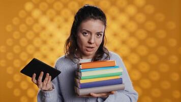 Woman debating between physical books and ebooks on ereader, isolated over studio background. Bookworm comparing paper novels and digital literature on tablet, technology concept, camera B video