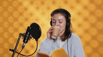 Voice actor reads book while enjoying coffee, recording audiobook using microphone, studio background. Woman using mic and headphones to produce digital recording of novel, drinking beverage, camera A video