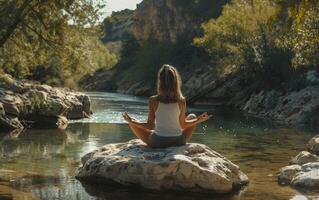 un mujer es sentado en un rock por un río, practicando yoga foto