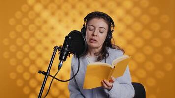 Narrator wearing headset reading aloud from book into mic against yellow background. Upbeat professional voice actor recording audiobook, creating engaging media content for listeners, camera B video