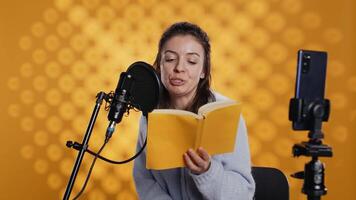 influenceur tournage en train de lire livre avec téléphone intelligent sur trépied, création livre audio, studio Contexte. femme vlogging avec mobile téléphone tandis que produisant numérique enregistrement sur roman en utilisant microphone, caméra b video