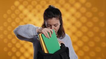 Portrait of woman rummaging through backpack, taking book out, isolated over studio background. Student removing textbook used for educational purposes from school rucksack, camera B video