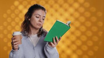 cansado mulher lendo livro, segurando copo do café, isolado sobre estúdio fundo. Exausta leitor ávido desfrutando romance e cafeína bebida, em falta energia, lutando para fique acordado, Câmera b video