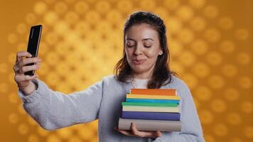 Reading enthusiast woman filming social media s with smartphone holding stack of books, studio background. Cheerful nerd with pile of textbooks in arms recording with selfie camera, camera B video