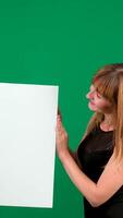 Smiling woman holding blank white banner and looking at it with friendly smile on green background vertical video