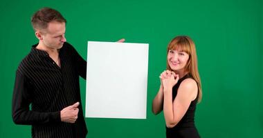 A guy and a girl in black clothes look at a white screen on a green background. I smile and point at the screen video