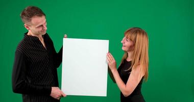 A guy and a girl in black clothes look at a white screen on a green background. I smile and point at the screen video