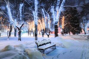 invierno parque a noche con Navidad decoraciones, luces, pavimento cubierto con nieve y arboles foto