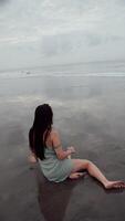 menina sentado em Preto molhado areia em a oceano, vertical video