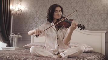 beautiful girl playing the violin while sitting on the bed at home, hand-held shooting video