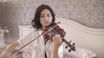 beautiful girl playing the violin while sitting on the bed at home, hand-held shooting video