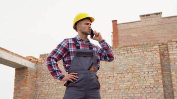 Construction worker in uniform and safety equipment have job on building. Talking by phone video