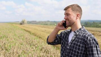 Farmer using a cell phone and holding notebook with a combine harvester in a wheat field on background video