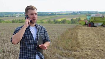 Farmer using a cell phone and holding notebook with a combine harvester in a wheat field on background video