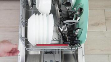 un hombre usos moderno aparato a mantener el hogar limpio. joven hombre cargando sucio platos dentro un lavavajillas máquina. de cerca, parte superior ver video
