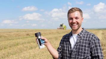 Young farmer inspects the state of wheat with special equipment. Combine harvester working in background video