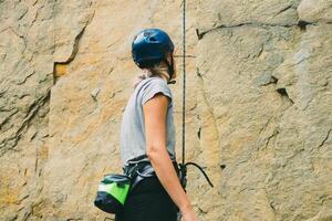 joven atlético mujer en equipo en pie en frente de Roca rock al aire libre y consiguiendo Listo a trepar. formación zona para al aire libre actividades. extremo deporte. posterior ver foto