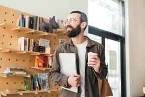 elegante y moderno barbado hombre en lentes sonriente y Bebiendo café en un café tienda en un soleado día, participación un ordenador portátil. persona de libre dedicación foto