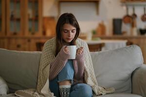 joven mujer vistiendo un de punto suéter sentado a hogar en el sofá envuelto en un tartán Bebiendo caliente café o té. congelado mujer será calentar arriba en un acogedor vivo habitación. foto