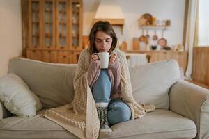 Woman freezes in winter time. Young girl wearing warm woolen socks and wrapped into plaid, holding a cup of hot drink while sitting on sofa at home. Keep warm. photo