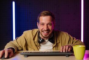 hermoso hombre en camisa ropa y auriculares sentado a un mesa en un habitación con neón luces y jugando juegos en el computadora con un sonrisa en su cara mirando a el pantalla. foto