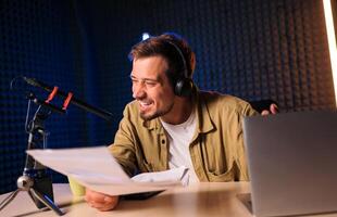 Smiling and gesturing radio host with headphones reading news from paper into studio microphone at radio station with neon lights photo