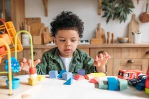 Concentrated little african american boy with bushy curly hair playing with colorful wooden blocks, enjoying table games playing alone at home kitchen , close up photo