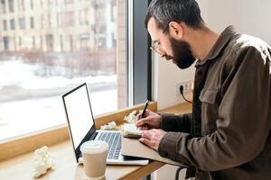 un caucásico hombre escribir en bloc en un cafetería. trabajo proceso. un hombre con un barba en un camisa es sentado en un café con un computadora portátil, remoto trabajar, Lanza libre foto