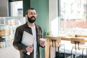 Smiling bearded creative man in glasses standing holding laptop and cup of coffee in cafe looking out window. Freelancer and modern interior photo