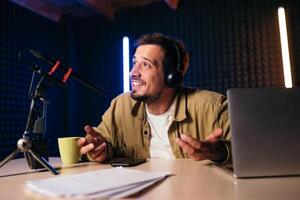 Young stylish man in mustard shirt with headphones gesturing at microphone and sharing story with audience while sitting at desk in studio with neon lighting and recording podcast photo