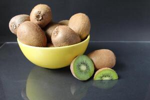 todavía vida de kiwi Fruta en un amarillo cuenco plato y piezas de jarra siguiente a en un negro antecedentes con un reflexión de allí es un copyspace foto