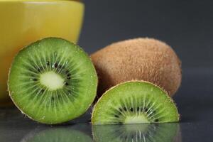 todavía vida de kiwi Fruta en un amarillo cuenco plato y piezas de jarra siguiente a en un negro antecedentes con un reflexión de allí es un copyspace foto