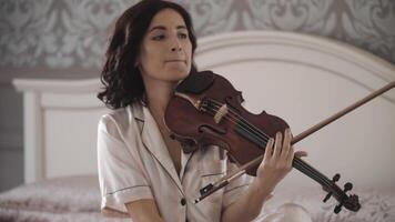 lindo menina jogando a violino enquanto sentado em a cama às lar, portátil tiroteio, fechar-se video